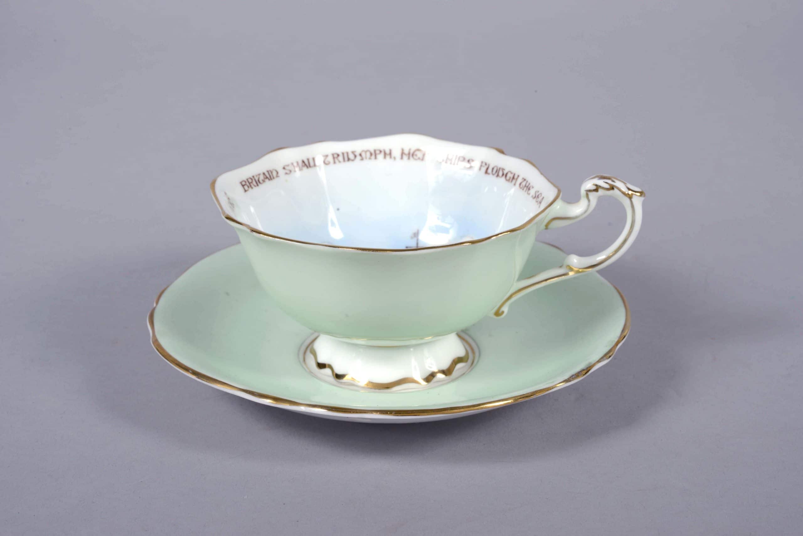 Green fine china cup with painting of three battleships. Words around the inside of the rim read: ‘Britain shall Triumph, her ships plough the sea, her standard be justice, her watchword be free’. Green fine china saucer with gold rim and Officers cap badge painted in the centre. This cup and saucer is part of the ‘Paragon Patriotic’ series produced during World War Two.