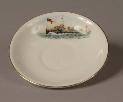Demi Tasse white porcelain saucer. Top of saucer is decorated with a picture of a World War One torpedo boat with a white ensign at the jack. Words on the saucer read: ‘H.M Torpedo Boat’.