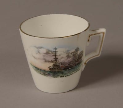 Demi Tasse white porcelain coffee cup with a gold rim around the top. Front of cup is decorated with a picture of a World War One battleship firing a broadside with painted words: ‘H.M Battleship in action’.